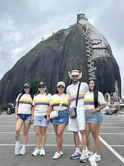 Small family group of tourists wearing matching shirts posing confidently at the base of the massive Piedra del Peñol rock in Guatapé.