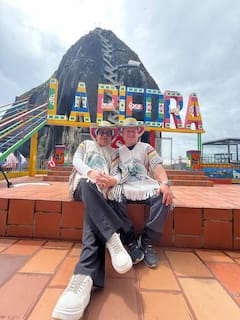 Two tourists wearing traditional Colombian ruanas and sombreros, posing for an iconic photo in front of the massive 'LA PIEDRA' sign at the base of the El Peñol rock.